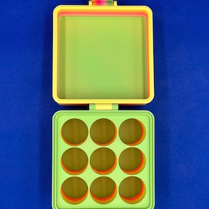 May include: A square, hinged storage container with a lime green interior. The top lid is open, revealing a plain surface, while the bottom section features a grid of nine circular compartments. The outer edges display a gradient of rainbow colors.