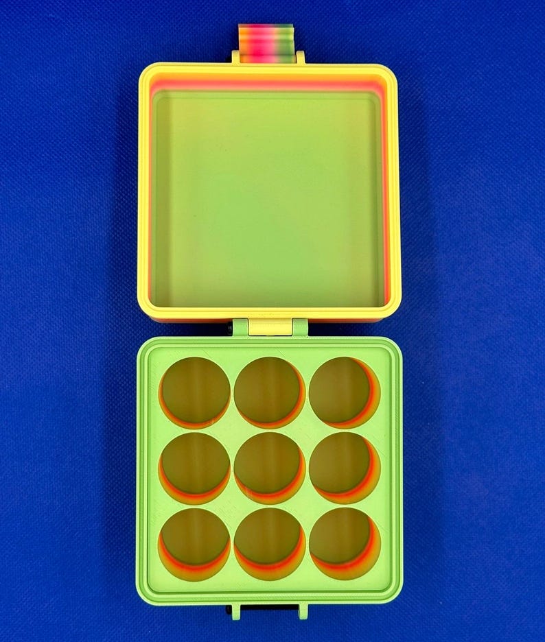 May include: A square, hinged storage container with a lime green interior. The top lid is open, revealing a plain surface, while the bottom section features a grid of nine circular compartments. The outer edges display a gradient of rainbow colors.