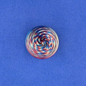 May include: A circular glass cabochon featuring a swirling pattern of red, white, and blue colors. The design radiates from the center in a spiral motion, creating a multi-colored effect with smooth, polished surfaces and a rounded dome shape.