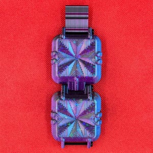 Puede incluir: Un objeto cuadrado de dos secciones con un acabado iridiscente en tonos violeta y azul, que presenta un patr&oacute;n geom&eacute;trico de estrella en cada cara. La estructura incluye un conector superior rectangular y crestas.