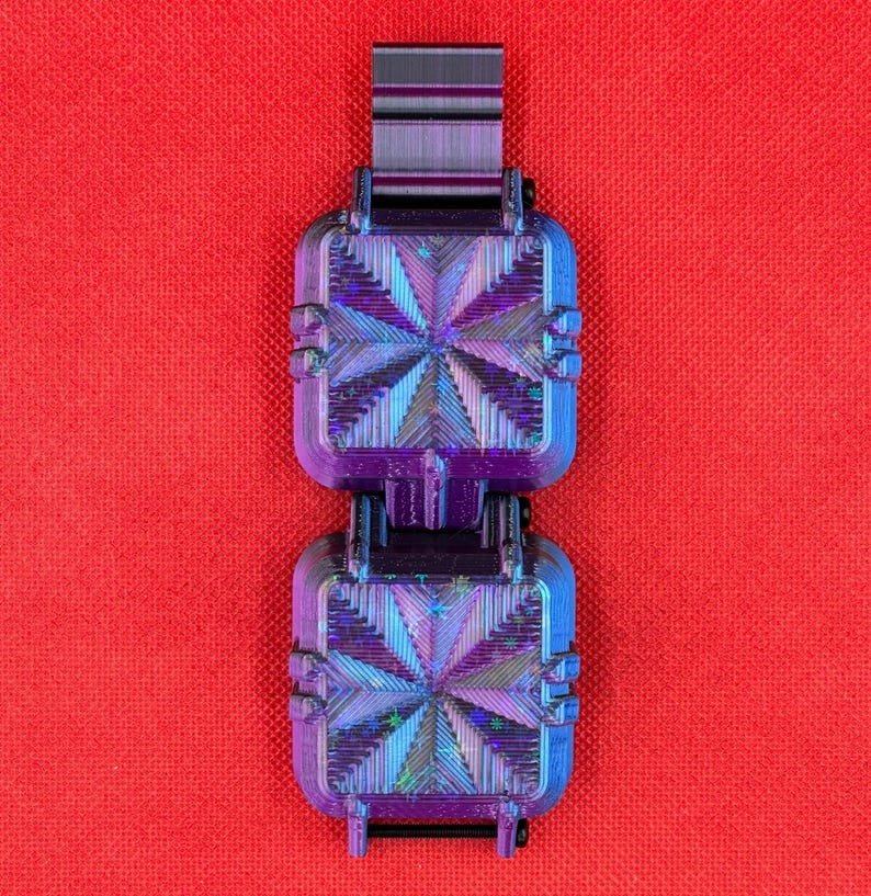 Puede incluir: Un objeto cuadrado de dos secciones con un acabado iridiscente en tonos violeta y azul, que presenta un patr&oacute;n geom&eacute;trico de estrella en cada cara. La estructura incluye un conector superior rectangular y crestas.