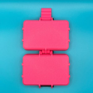 Puede incluir: Un estuche de almacenamiento rectangular de pl&aacute;stico en color rosa intenso con tapa abatible y mecanismo de cierre seguro. El estuche presenta una superficie texturizada y se muestra en posici&oacute;n abierta, revelando dos compartimentos id&eacute;nticos.