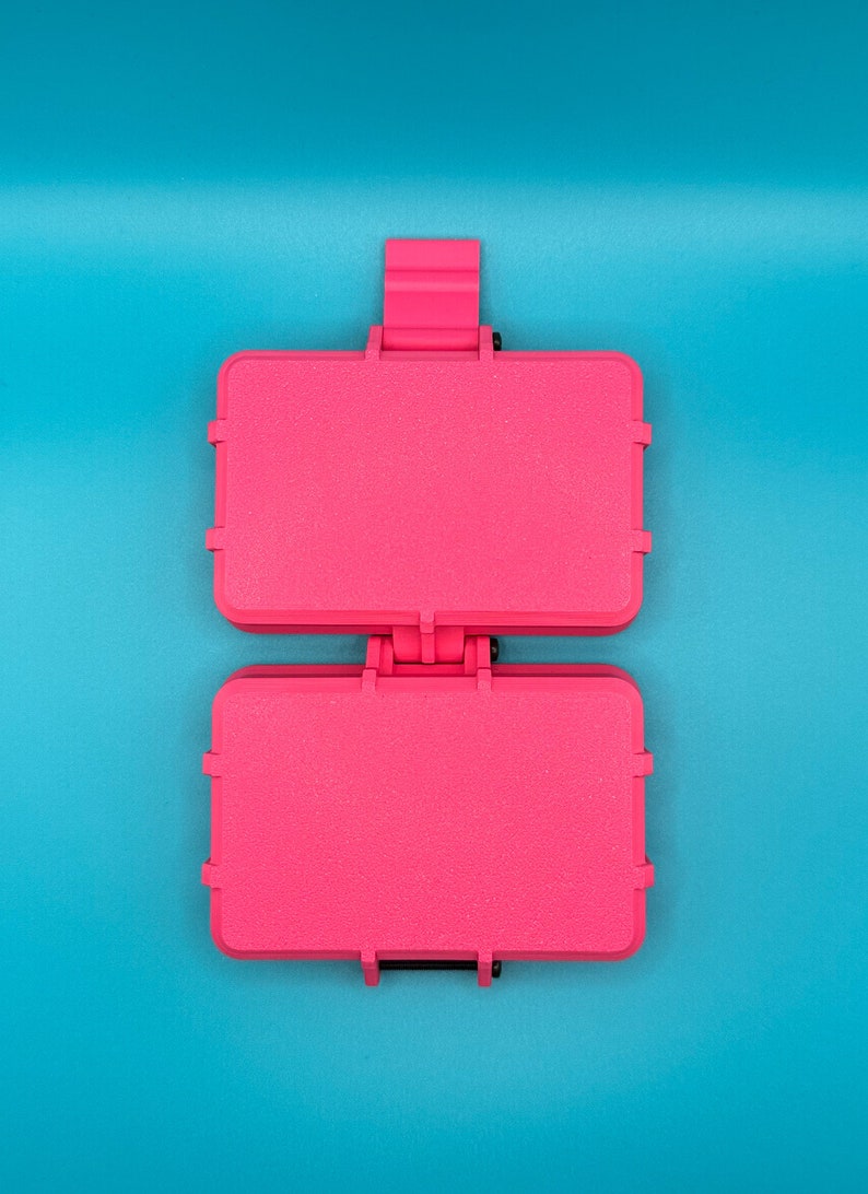 Puede incluir: Un estuche de almacenamiento rectangular de pl&aacute;stico en color rosa intenso con tapa abatible y mecanismo de cierre seguro. El estuche presenta una superficie texturizada y se muestra en posici&oacute;n abierta, revelando dos compartimentos id&eacute;nticos.