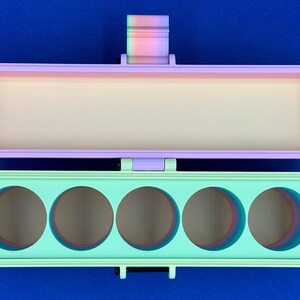 May include: A rectangular hinged storage case with a light purple lid and a mint green base. The base features five circular cutouts, and the hinge mechanism displays a gradient of rainbow colors. The interior of the case is a solid cream color.