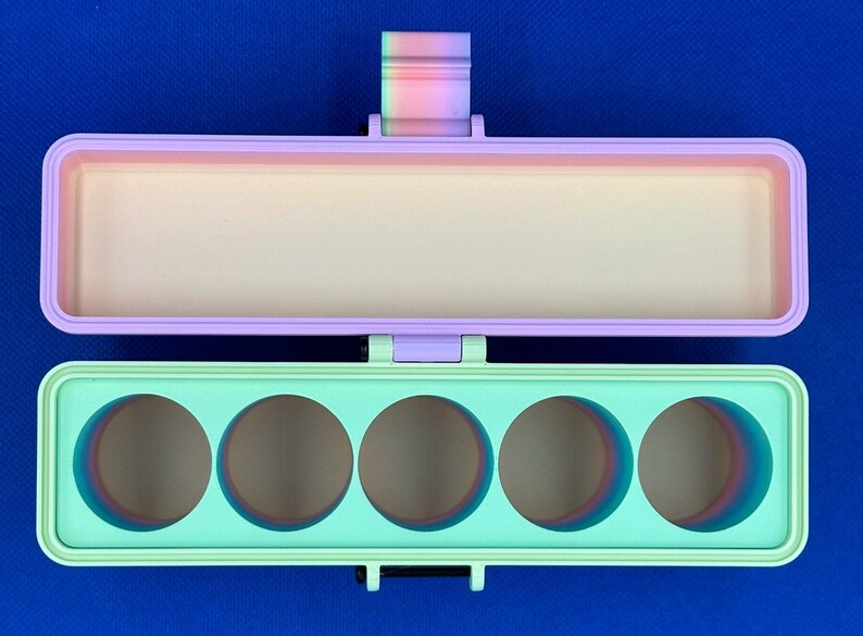 May include: A rectangular hinged storage case with a light purple lid and a mint green base. The base features five circular cutouts, and the hinge mechanism displays a gradient of rainbow colors. The interior of the case is a solid cream color.