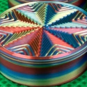May include: A round, multi-colored container featuring a vibrant, radial star pattern on its lid. The sides of the container display horizontal bands of shifting colors, including blue, red, and yellow, creating a gradient effect across the surface.