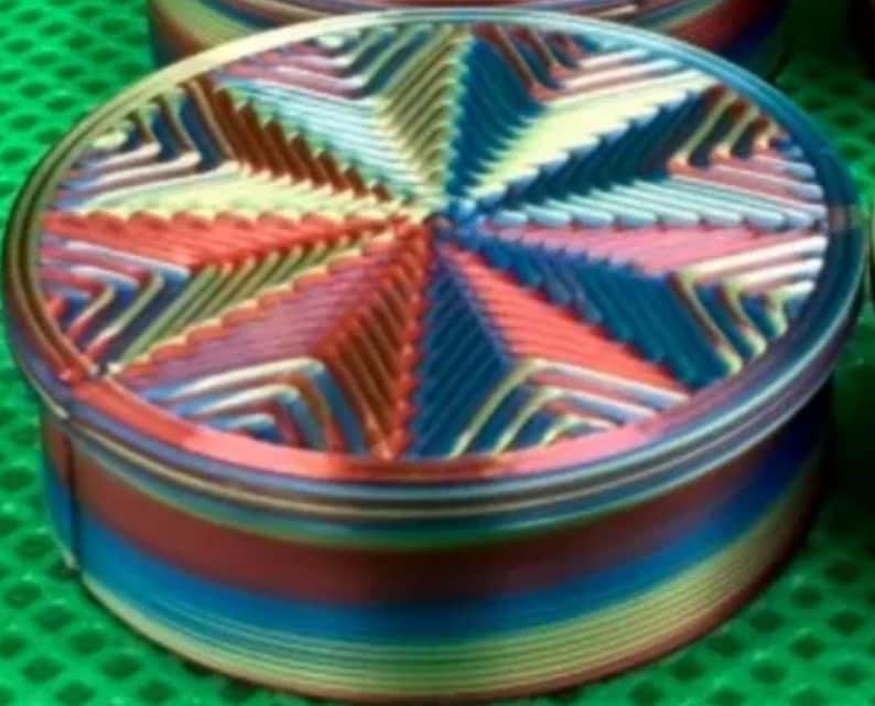 May include: A round, multi-colored container featuring a vibrant, radial star pattern on its lid. The sides of the container display horizontal bands of shifting colors, including blue, red, and yellow, creating a gradient effect across the surface.