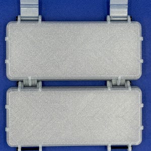 May include: A pair of rectangular, silver-colored storage cases with a textured finish. Each case features a hinged lid, reinforced edges, and two vertical mounting clips on the top edge. The two cases are connected by a central hinge mechanism.