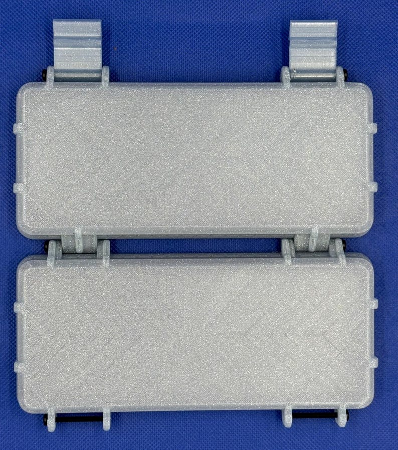 May include: A pair of rectangular, silver-colored storage cases with a textured finish. Each case features a hinged lid, reinforced edges, and two vertical mounting clips on the top edge. The two cases are connected by a central hinge mechanism.