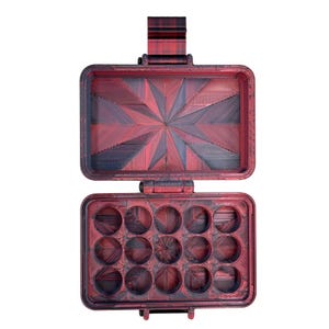 Puede incluir: Un estuche de almacenamiento rectangular con tapa abatible, con un acabado degradado en rojo oscuro y negro. La tapa interior muestra un patr&oacute;n geom&eacute;trico de estrella, mientras que la base contiene doce compartimentos circulares.