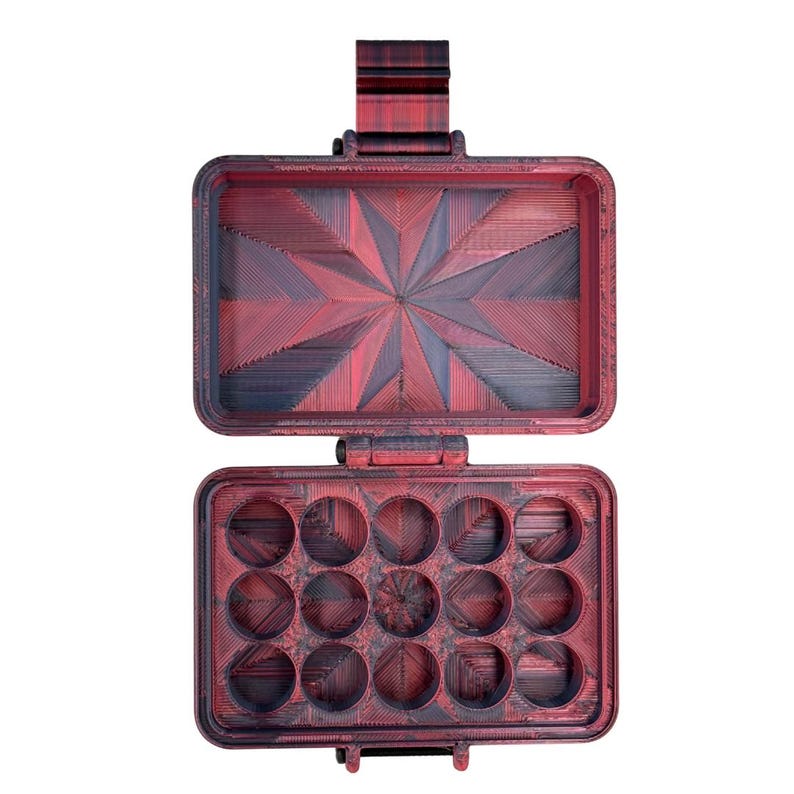 Puede incluir: Un estuche de almacenamiento rectangular con tapa abatible, con un acabado degradado en rojo oscuro y negro. La tapa interior muestra un patr&oacute;n geom&eacute;trico de estrella, mientras que la base contiene doce compartimentos circulares.
