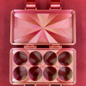 May include: A red plastic storage box with a hinged lid and six round compartments. The box has a geometric pattern on the lid and the compartments.