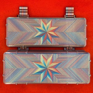 May include: A rectangular, hinged storage case featuring an iridescent, multi-coloured starburst pattern on its surface. The case has two small, striped fabric tabs attached to the top edge, and the overall design displays a geometric, radiating texture.
