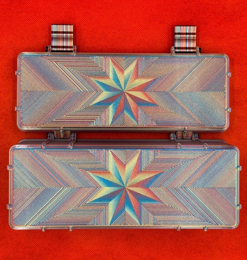 May include: A rectangular, hinged storage case featuring an iridescent, multi-coloured starburst pattern on its surface. The case has two small, striped fabric tabs attached to the top edge, and the overall design displays a geometric, radiating texture.