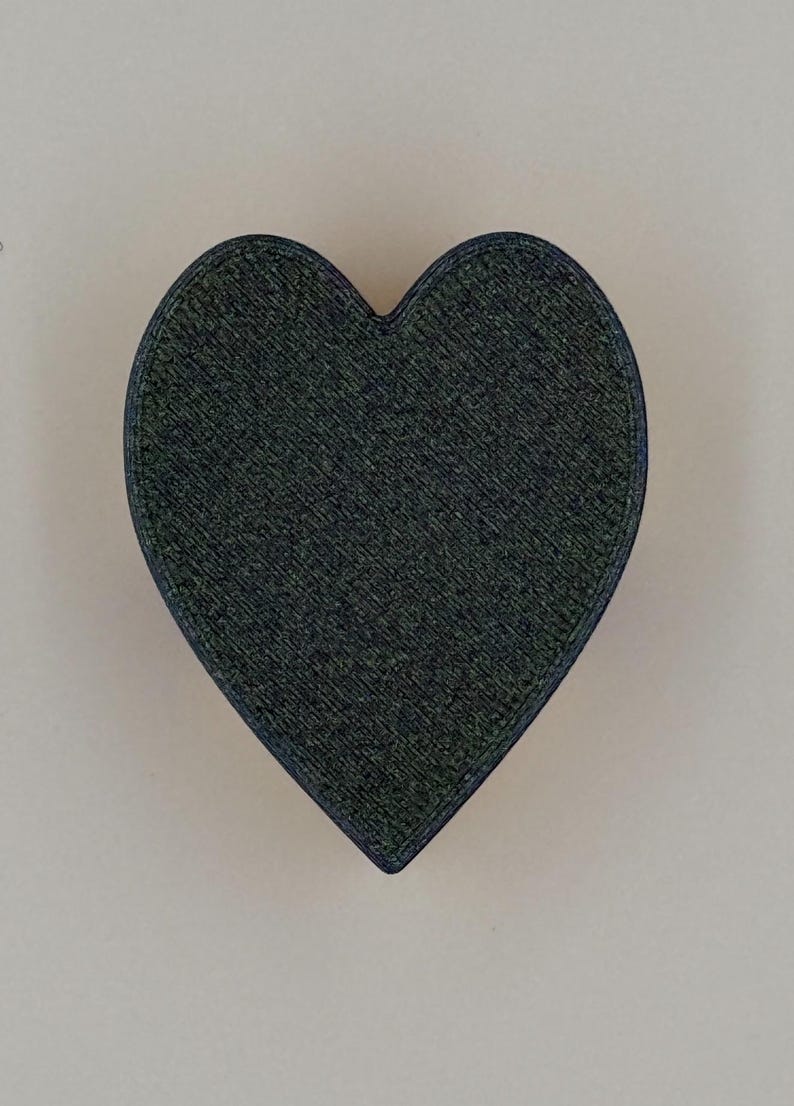 May include: A dark green heart-shaped object with a textured surface and a dark blue outline. The heart is centered against a light beige background. The object appears to be made of a solid material.