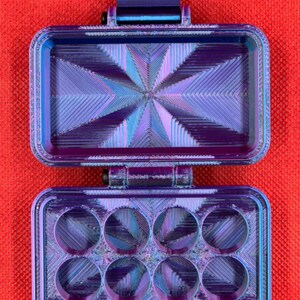 May include: A small, purple and blue, six-compartment storage case with a hinged lid. The lid has a textured, geometric design.