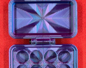 May include: A rectangular, iridescent purple and blue hinged storage case with a textured, geometric starburst pattern on the interior surfaces. The bottom section features eight circular cutouts arranged in two rows of four, while the top lid is hollow.