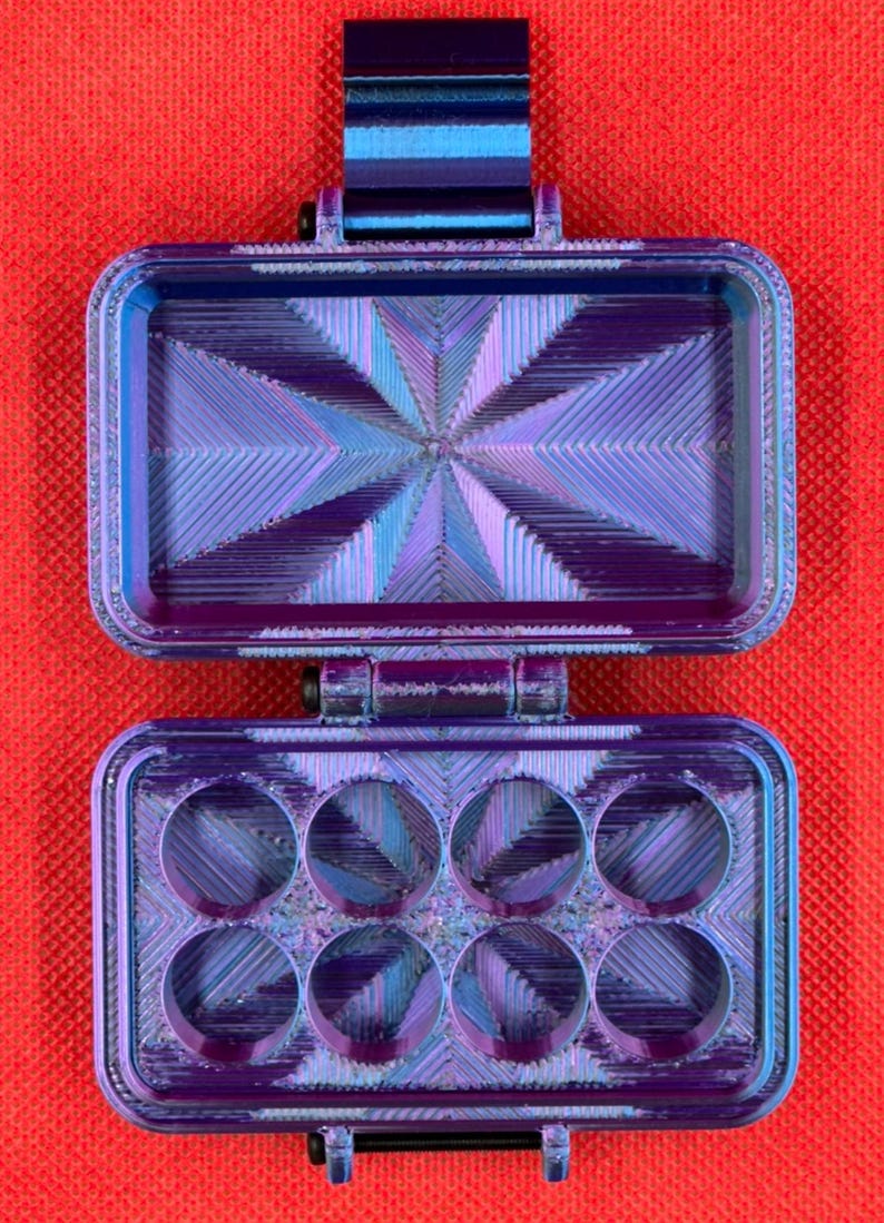 May include: A rectangular, iridescent purple and blue hinged storage case with a textured, geometric starburst pattern on the interior surfaces. The bottom section features eight circular cutouts arranged in two rows of four, while the top lid is hollow.