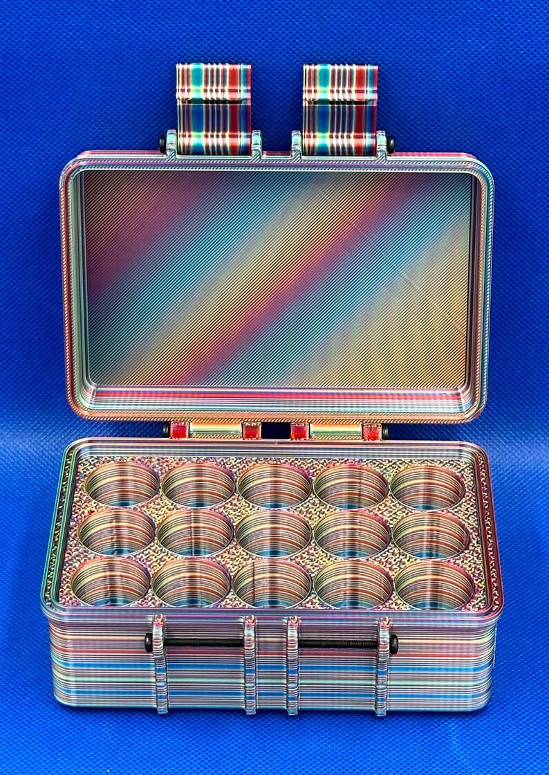May include: A rectangular storage case with an open lid, featuring a multi-colored iridescent finish with horizontal and diagonal striped patterns. The interior base contains fifteen circular compartments arranged in three rows of five.