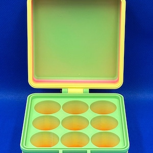 Puede incluir: Un contenedor de almacenamiento cuadrado con bisagra que presenta un degradado de colores rosa, naranja, amarillo y verde. La base interior contiene nueve compartimentos circulares en una cuadrícula de tres por tres.