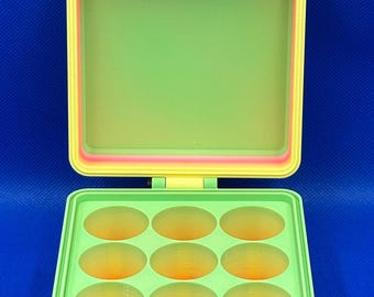 May include: A square, hinged storage container featuring a gradient color scheme of pink, orange, yellow, and green. The interior base contains nine circular compartments arranged in a three-by-three grid, while the lid remains open.