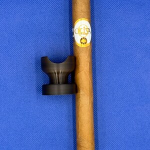 May include: A brown cigar with a gold band that says "OLIVA" on a blue surface with a black cigar rest.