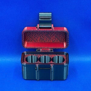 May include: A rectangular storage case with a hinged lid, featuring a black exterior and a red interior with a textured pattern. The base contains three circular compartments arranged in a row, and a black latch secures the lid.