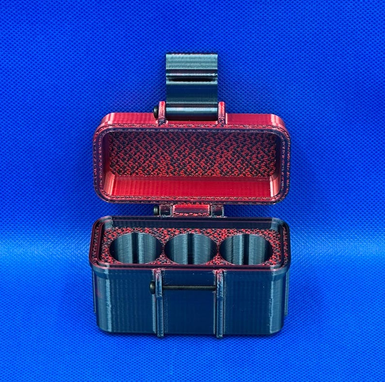 May include: A rectangular storage case with a hinged lid, featuring a black exterior and a red interior with a textured pattern. The base contains three circular compartments arranged in a row, and a black latch secures the lid.