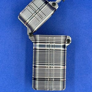 May include: A rectangular metal lighter with a hinged lid in an open position. The surface features a metallic finish with a repeating pattern of horizontal and vertical lines in shades of silver, gray, and black. Two small black screws are visible.