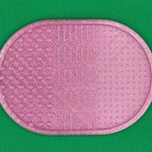 May include: An oval-shaped pink makeup brush cleaning mat featuring a textured surface with three distinct sections. The left side has raised circular bumps, the center has vertical ridges, and the right side has a fine dotted pattern.