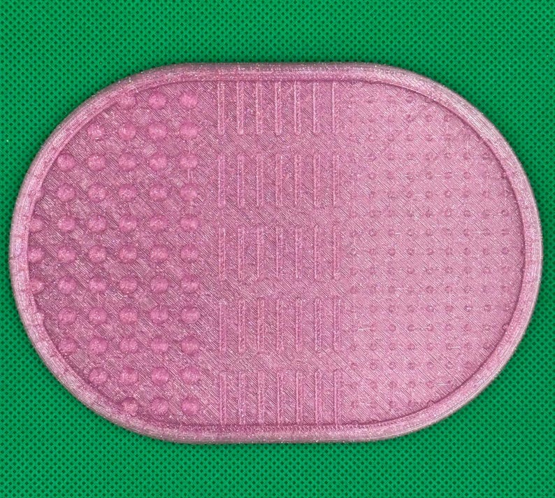 May include: An oval-shaped pink makeup brush cleaning mat featuring a textured surface with three distinct sections. The left side has raised circular bumps, the center has vertical ridges, and the right side has a fine dotted pattern.