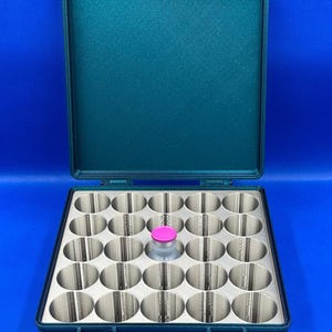 May include: A teal-colored plastic case with a hinged lid, containing a foam insert with 24 circular compartments. One compartment contains a small glass vial with a pink cap.
