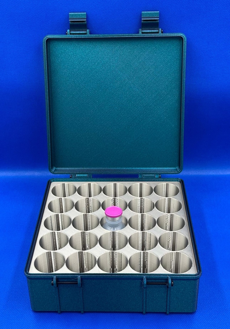 May include: A teal-colored plastic case with a hinged lid, containing a foam insert with 24 circular compartments. One compartment contains a small glass vial with a pink cap.