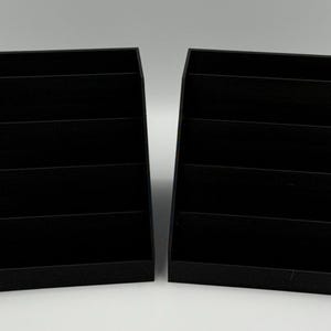 May include: Two black, tiered display shelves. Each shelf has four horizontal compartments. The shelves are made of a matte black material and are set against a white background. The shelves are designed to hold and display items.