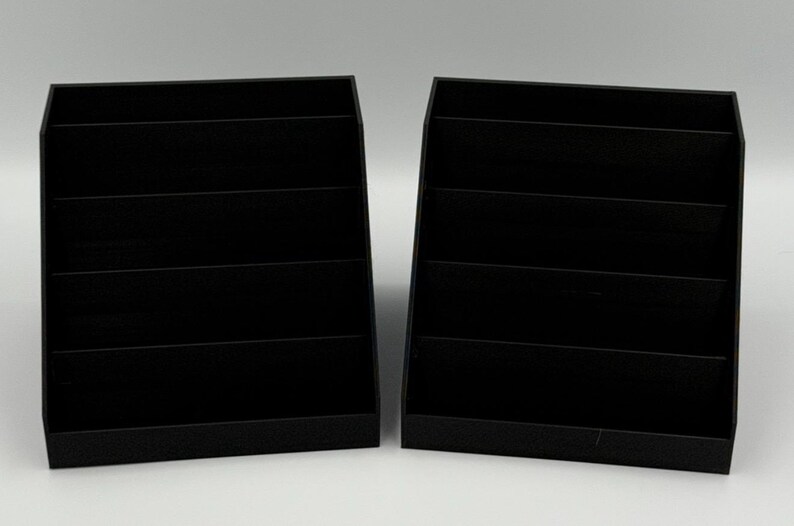 May include: Two black, tiered display shelves. Each shelf has four horizontal compartments. The shelves are made of a matte black material and are set against a white background. The shelves are designed to hold and display items.