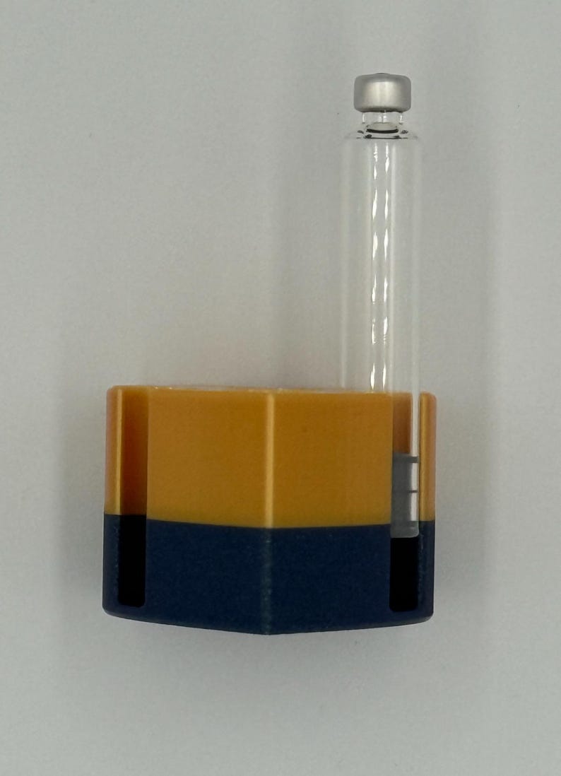 May include: A clear glass vial with a silver cap is attached to a two-tone container. The container is a combination of yellow and navy blue. The vial is cylindrical and the container is rectangular.
