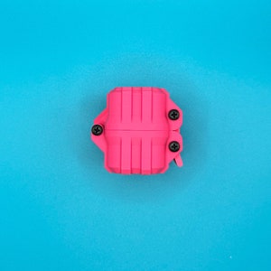 May include: A bright pink, rectangular, ribbed plastic case featuring three visible black Phillips head screws securing the two halves together. The object has a structured, industrial appearance with vertical grooves along its surface.
