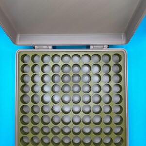 May include: A gray plastic storage case with a green foam insert. The insert has 64 round compartments.
