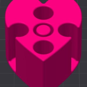 Puede incluir: Un objeto rosa brillante en forma de corazón con una base cilíndrica. La parte superior presenta tres agujeros circulares y una hendidura circular central más pequeña. El objeto tiene una apariencia suave y sólida sobre un fondo gris oscuro.