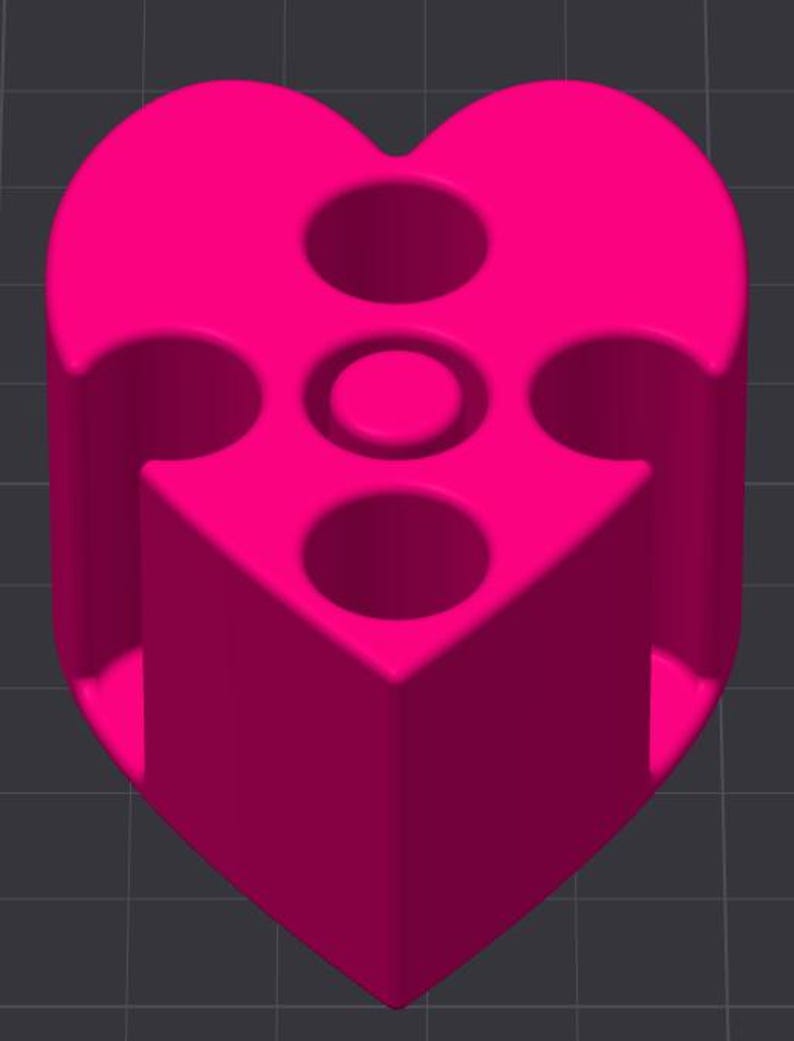 May include: A bright pink, heart-shaped object with a cylindrical base. The top features three circular holes and a central, smaller circular indentation. The object has a smooth, solid appearance and is set against a dark gray background.