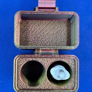 May include: A rectangular, open storage case with a metallic, iridescent finish featuring a textured, cross-hatched pattern. The interior base contains two circular compartments, one of which holds a small white plastic pump dispenser.