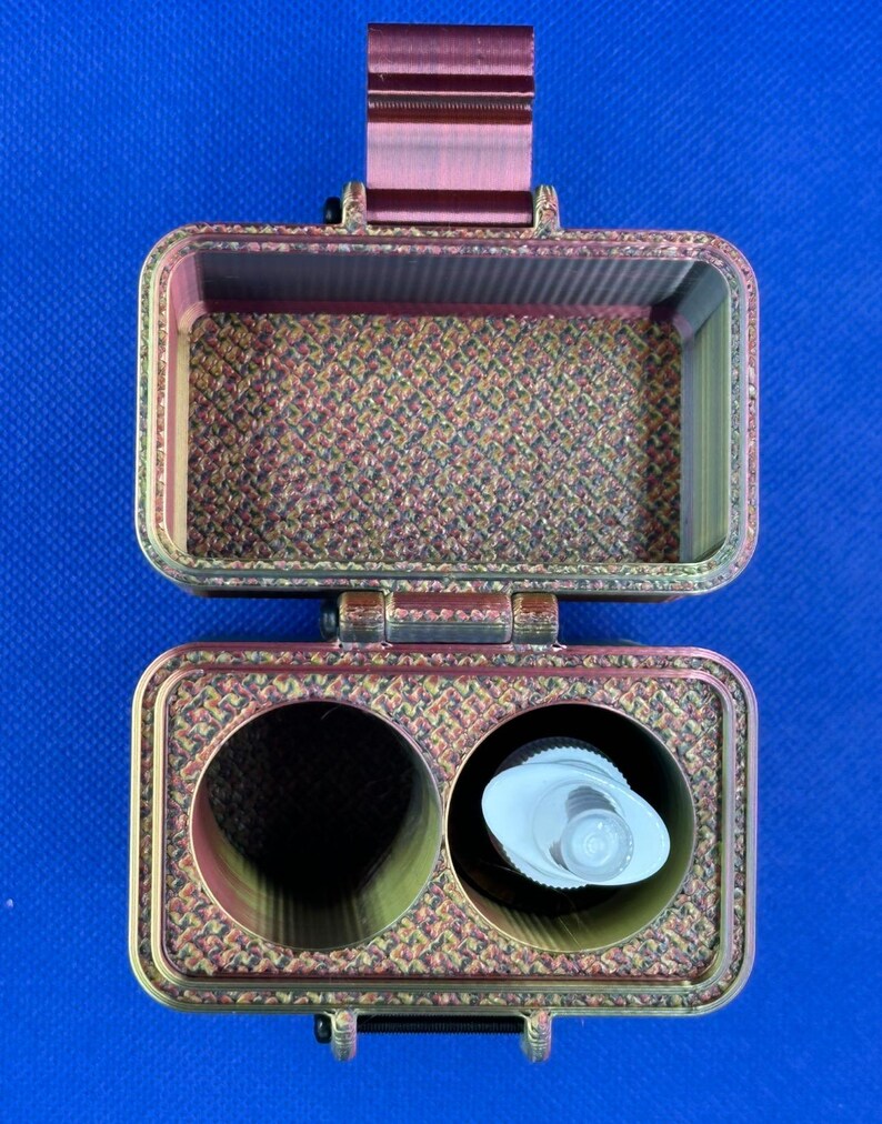 May include: A rectangular, open storage case with a metallic, iridescent finish featuring a textured, cross-hatched pattern. The interior base contains two circular compartments, one of which holds a small white plastic pump dispenser.