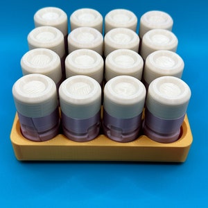 May include: A set of sixteen cylindrical containers with light purple bases and off-white lids, arranged in a four-by-four grid within a rectangular yellow tray. Each lid features a textured, circular top surface with visible 3D print lines.