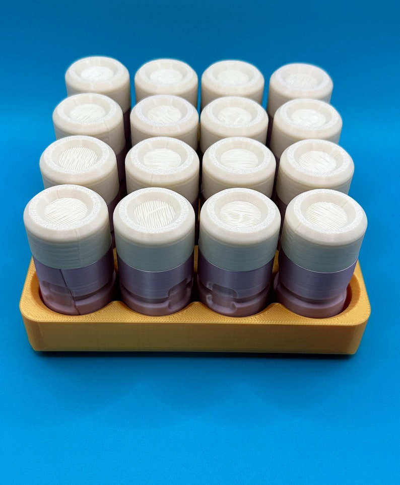 May include: A set of sixteen cylindrical containers with light purple bases and off-white lids, arranged in a four-by-four grid within a rectangular yellow tray. Each lid features a textured, circular top surface with visible 3D print lines.