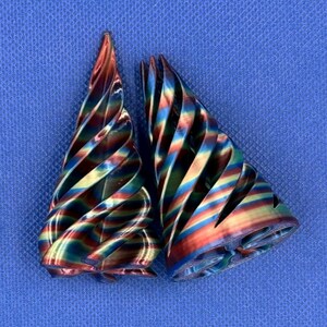May include: Two conical objects with a spiraling, twisted design. The surfaces feature a metallic, iridescent gradient transitioning between shades of red, gold, blue, and green. The structures are hollow with open, layered, and ribbed textures.