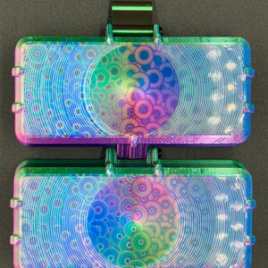May include: A rectangular, dual-compartment case featuring a vibrant, iridescent finish that shifts between shades of green, blue, and purple. The surface is textured with a repeating pattern of concentric circles and a metallic clip.