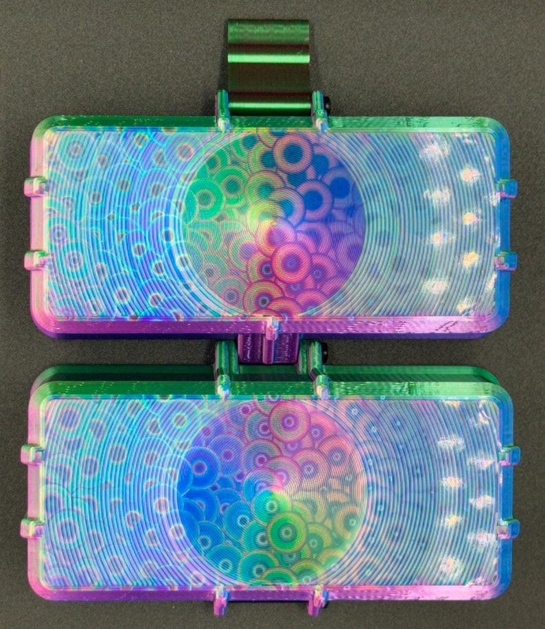 May include: A rectangular, dual-compartment case featuring a vibrant, iridescent finish that shifts between shades of green, blue, and purple. The surface is textured with a repeating pattern of concentric circles and a metallic clip.