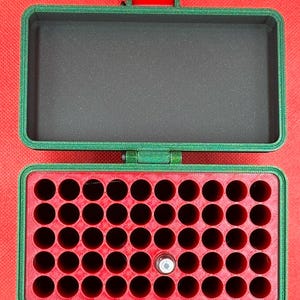 May include: A rectangular green storage case with a hinged lid and a red latch. The interior features a red insert with forty-eight circular holes for small items, and one small silver cylindrical object is placed in one of the holes.