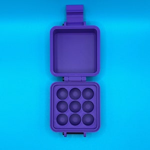 May include: A purple, square, hinged storage case with a latch. The top section is an open, empty compartment, while the bottom section features a grid of nine circular, concave indentations arranged in a three-by-three pattern.