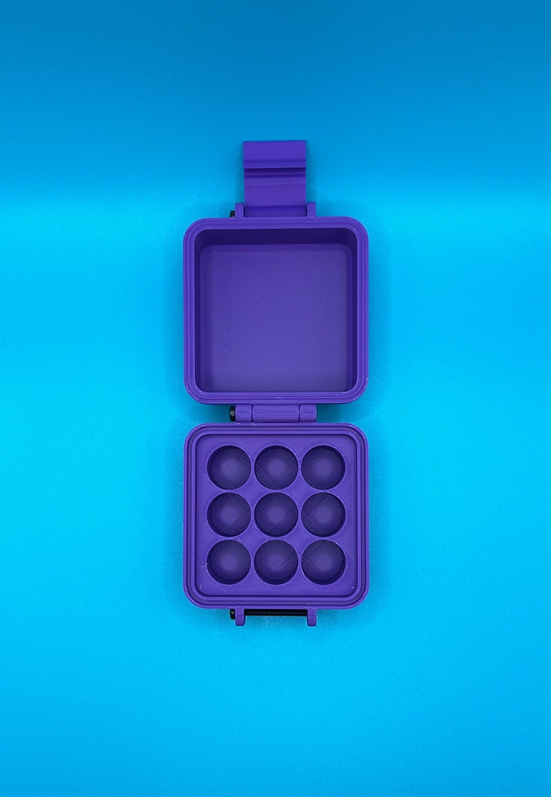 May include: A purple, square, hinged storage case with a latch. The top section is an open, empty compartment, while the bottom section features a grid of nine circular, concave indentations arranged in a three-by-three pattern.